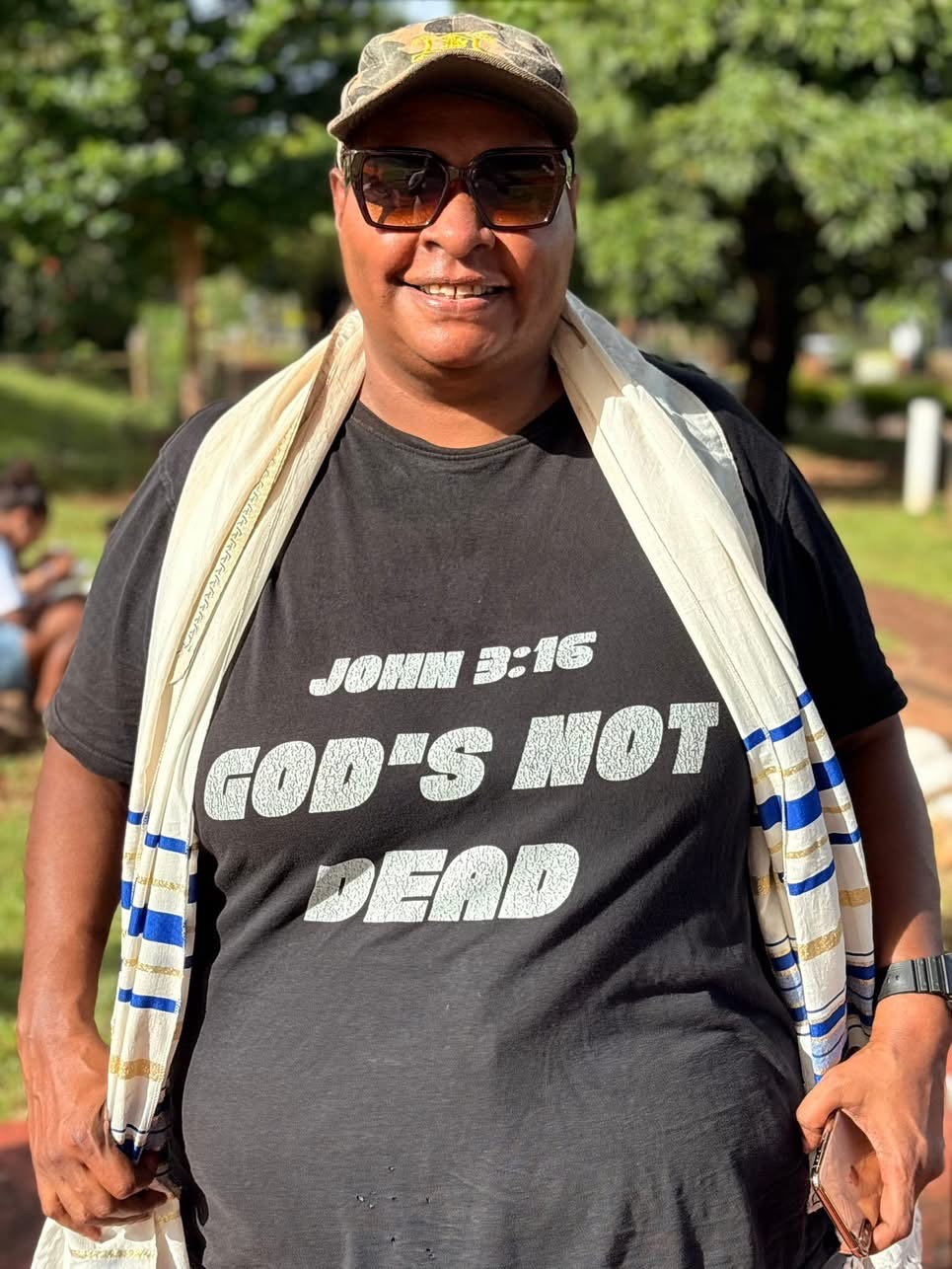 A person standing outdoors wearing a black T-shirt with a Christian message